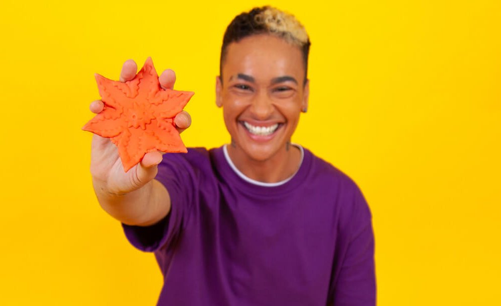 A smiling person against a yellow background is holding up a Coral Cute Little Fuckers Starsi to the camera. Their fingers comfortably fit in-between each one of the prongs of the design, and the Starsi is larger than their palm's size. | Kinkly Shop